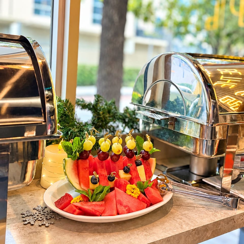 Buffet platter of watermelon wedges and fruit skewers beside polished stainless-steel serving chafers.