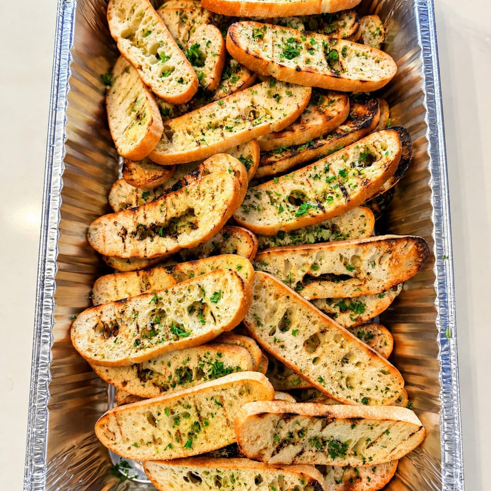 Aluminum tray filled with toasted garlic bread slices topped with herbs and browned, charred edges.