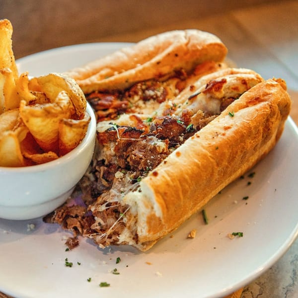 A cheesy steak sandwich on a white plate with a side of crispy potato chips.