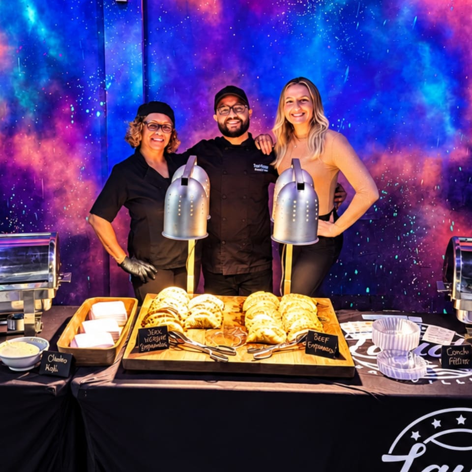 Three smiling caterers pose behind a buffet table serving empanadas against a colorful galaxy-themed backdrop.