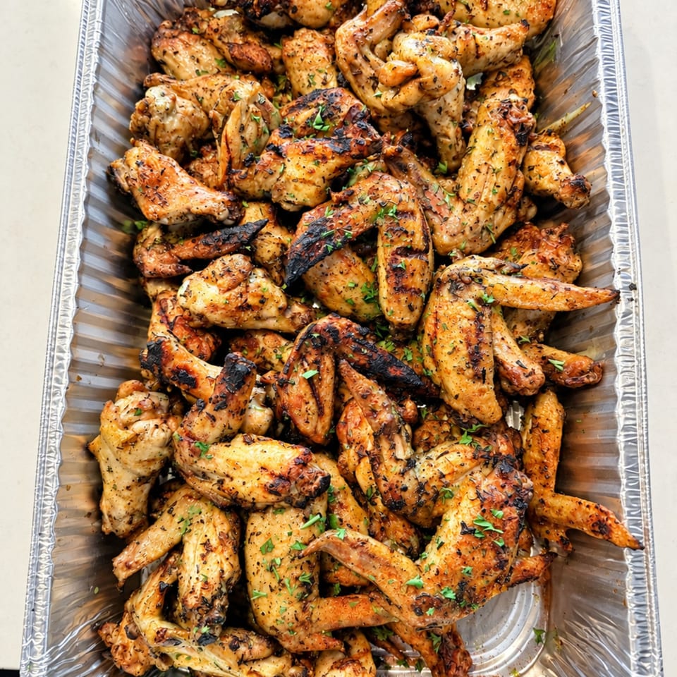 Aluminum tray filled with grilled, seasoned chicken wings garnished with chopped herbs.